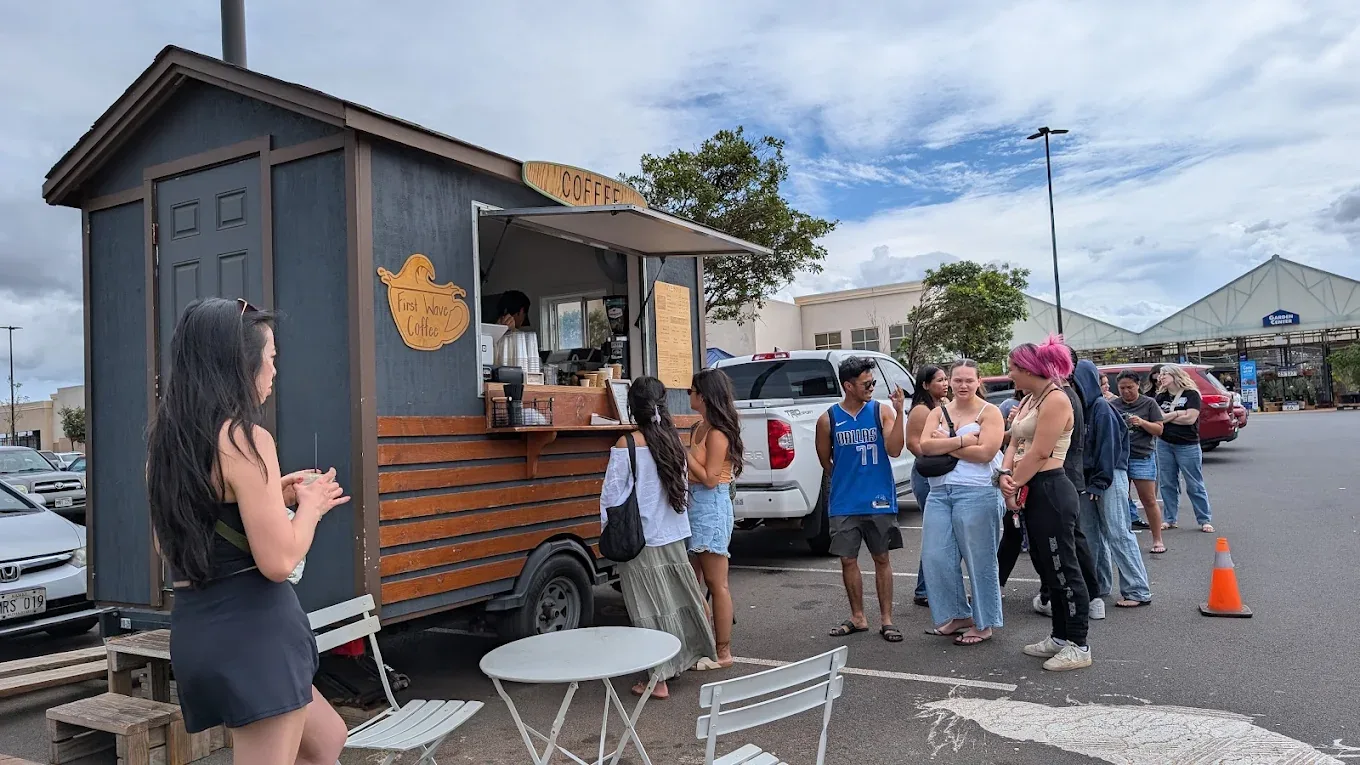 A line of customers waiting at the First Wave Coffee trailer in a sunny parking lot.