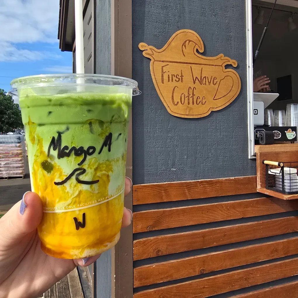 A layered mango matcha drink held up in front of the First Wave Coffee trailer sign.