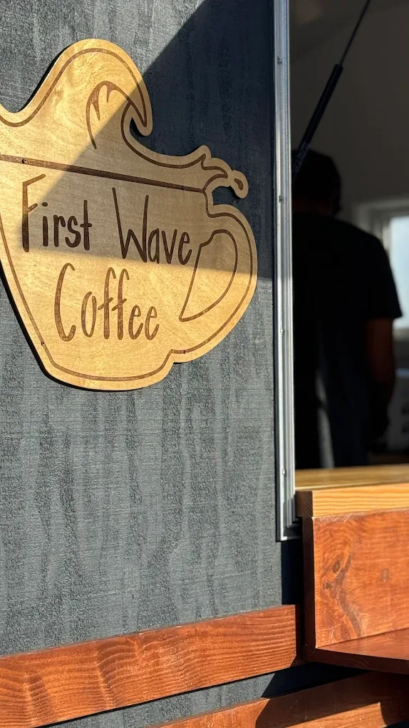 First Wave Coffee's hand-carved wooden cup-and-wave logo glowing in golden-hour light on the side of the trailer.
